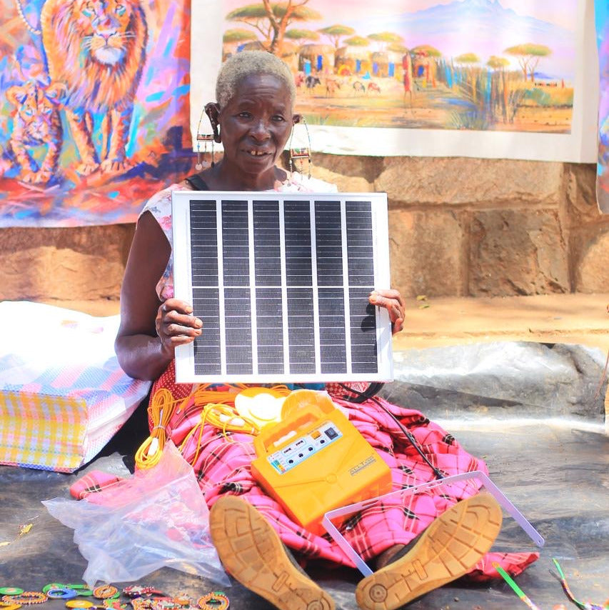 A woman sitting on the ground at what appears to be a market or craft fair holds up a small solar panel. She is wearing a pink patterned skirt and brown shoes, with a yellow electronic device resting on her lap.